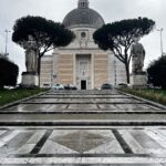 Facciata della Basilica dei Santi Pietro e Paolo all’Eur in una giornata nuvolosa, con la scalinata bagnata dalla pioggia e due statue ai lati dell’ingresso.