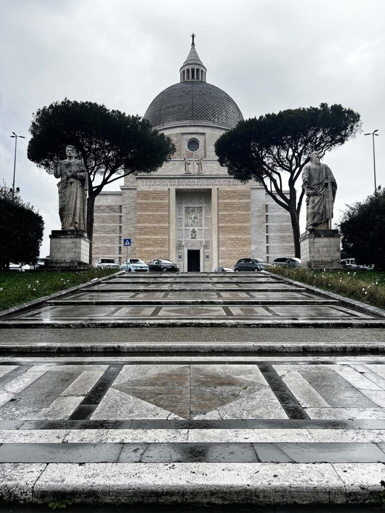 Facciata della Basilica dei Santi Pietro e Paolo all’Eur in una giornata nuvolosa, con la scalinata bagnata dalla pioggia e due statue ai lati dell’ingresso.