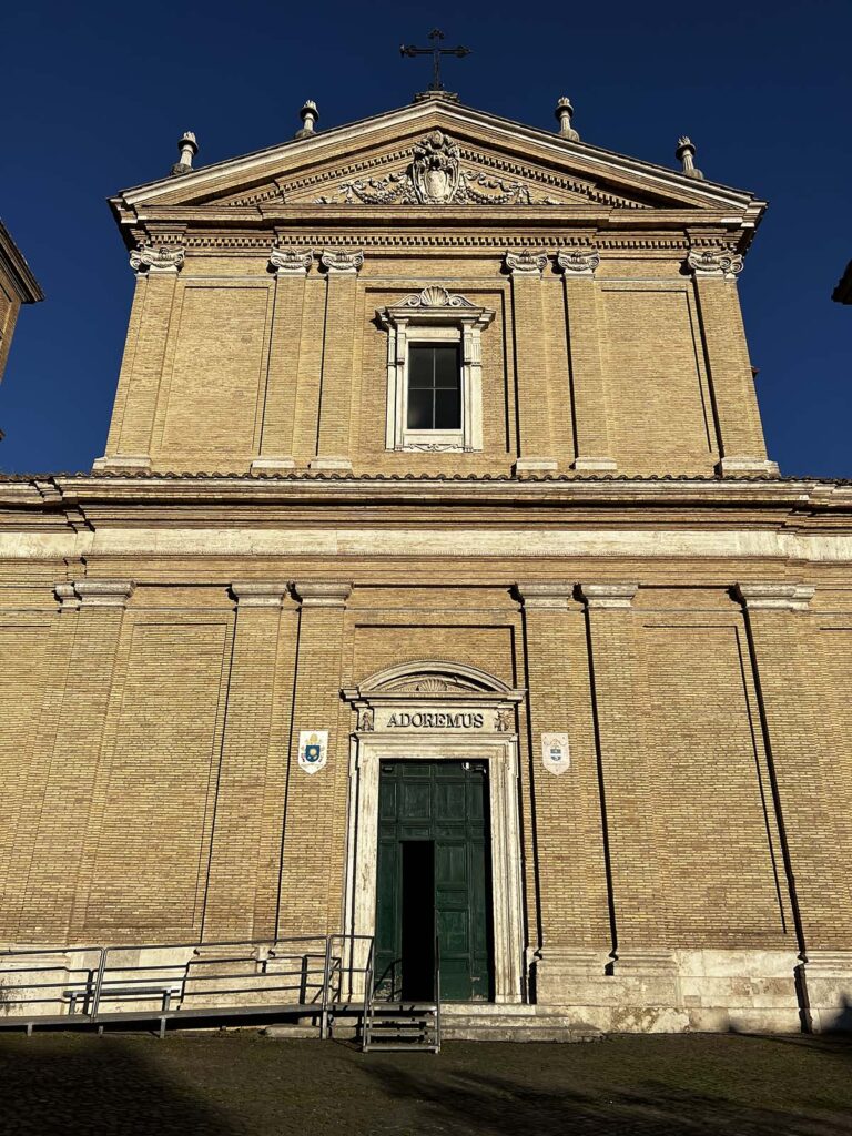 Facciata della Basilica di Sant’Anastasia al Palatino a Roma, illuminata dal sole del mattino, con la scritta “Adoremus” sopra il portale principale.