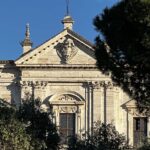 Facciata della chiesa San Gregorio al Celio illuminata dal sole, con una croce in cima e alberi che incorniciano la scena.