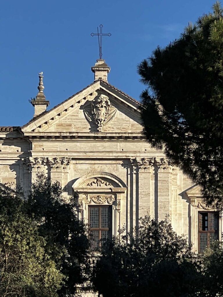Facciata della chiesa San Gregorio al Celio illuminata dal sole, con una croce in cima e alberi che incorniciano la scena.