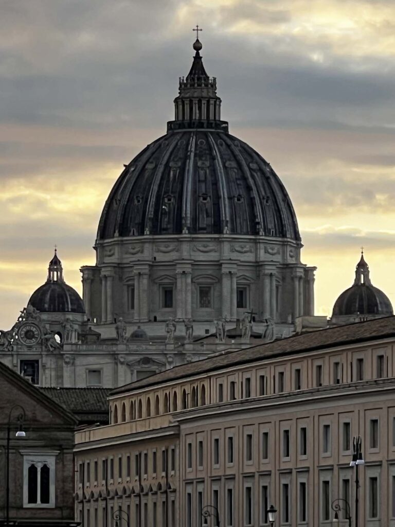 Cupola della Basilica di San Pietro illuminata dalla luce del tramonto con edifici in primo piano.